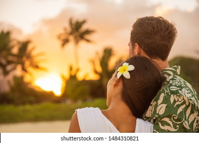 Romantic couple relaxing watching sunset on beach stroll view from back. Woman resting head on lover's shoulder on honeymoon vacation travel in summer Hawaii destination. Newlyweds people.