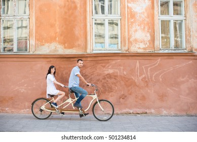 Romantic Couple Riding On Tandem Bike At The Street City Against The Background Of The Old Orange Wall With Windows. The Man Drives A Bicycle