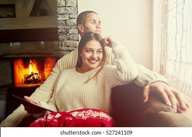 Romantic Couple Sharing A Digital Tablet Near A Wood Stove On A Winter Evening, Enjoying The Warm Christmas Atmosphere In Their Living Room