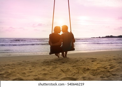 Romantic Couple Is Sitting And Kissing On Sea Beach On Rope Swing . Family Vacation On Honeymoon. Love And Relationship