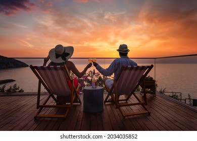 A romantic couple sitting in lounge chairs on summer vacation enjoys the sunset over the mediterranean sea with a glass of wine