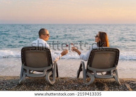 A romantic couple sitting in lounge chairs on summer vacation enjoys the sunset near sea with a glass of cocktail. Couple with refreshing cocktails relaxing in lounge chairs on beach