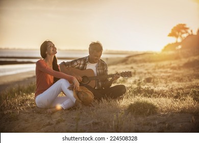 Romantic couple sitting on the beach at sunset with the man playing the guitar - Powered by Shutterstock