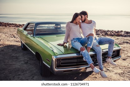 Romantic couple is sitting on green retro car on the beach. Handsome bearded man and attractive young woman with vintage classic car. Love story.