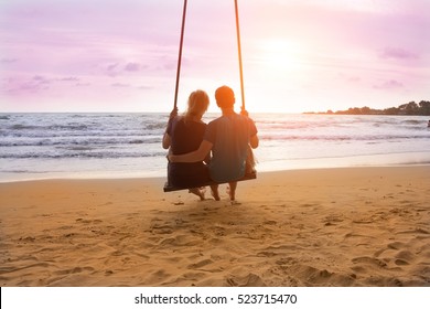 Romantic Couple Is Sitting On Sea Beach On Rope Swing And Looking At Sunset Horizon. Family Vacation On Honeymoon