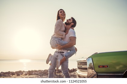 Romantic Couple Is Standing Near Green Retro Car On The Beach. Handsome Bearded Man And Attractive Young Woman With Vintage Classic Car. Love Story.Muscle Car