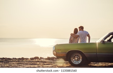 Romantic couple is standing near green retro car on the beach. Handsome bearded man and attractive young woman with vintage classic car. Love story.