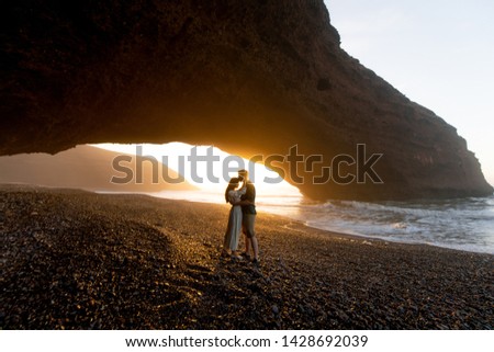 Romantic couple standing near natural huge arch on the beach. Handsome man and attractive woman enjoying sunset in Morocco.