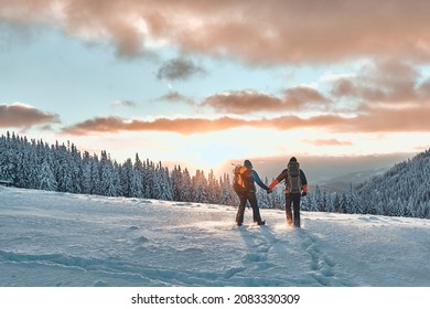 Romantic couple of tourists dressed in warm winter sportswear with tourist backpacks walking with trekking poles in the snowy pine mountains in an incredible sunset. Family, rest.