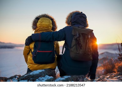 Romantic Couple Of Tourists Dressed In Warm Winter Sportswear With Tourist Backpacks Sitting In The Snowy Mountains In An Incredible Sunset. Ogoi Island, Baikal Lake. Family, Rest.