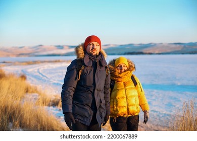 Romantic Couple Of Tourists Dressed In Warm Winter Sportswear With Tourist Backpacks Walking In The Snowy Mountains In An Incredible Sunset. Ogoi Island, Baikal Lake. Family, Rest.