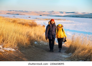 Romantic Couple Of Tourists Dressed In Warm Winter Sportswear With Tourist Backpacks Walking In The Snowy Mountains In An Incredible Sunset. Ogoi Island, Baikal Lake. Family, Rest.