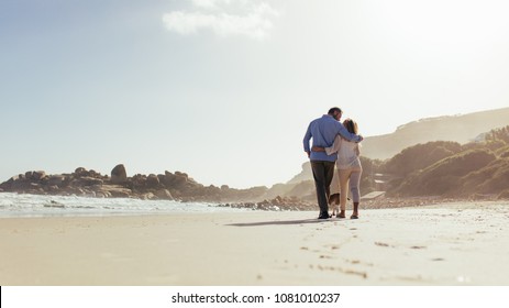 Romantic Couple Walking Along The Beach With A Dog. Loving Mature Couple Strolling On Sea Shore With Dog.