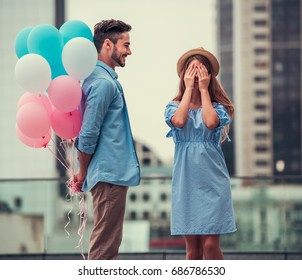 Romantic couple walking in the city. Girl is covering her eyes waiting for surprise, guy is holding balloons behind his back - Powered by Shutterstock