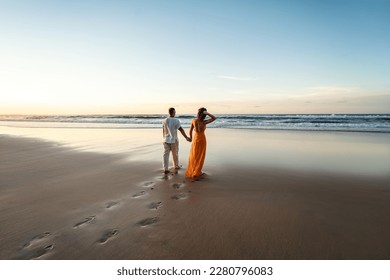 Romantic couple walking on sunset beach, enjoying evening light, relaxing on tropical summer vacation. Honeymoon. Love. Back view. Woman wearing orange maxi dress, man in linen pant and white shirt.
 - Powered by Shutterstock