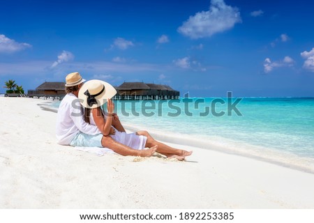 A romantic couple in white summer clothes sits hugging in the fine sand of a tropical beach in the Maldives