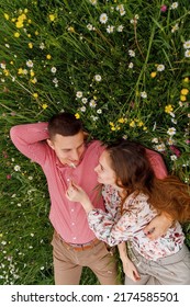 Romantic Couple Of Young People Lying Down On Grass In Field Of Spring Flowers. Woman Laying On The Shoulders Of Man And Looking Happily. View From Above.