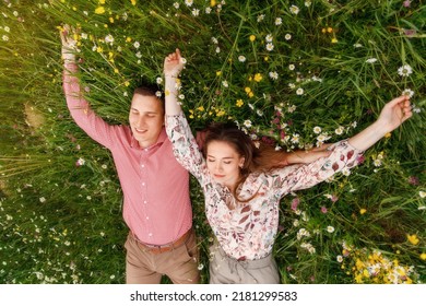 Romantic couple of young people lying down on grass in field of spring flowers. Woman laying on the shoulders of man and looking happily. View from above.