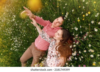 Romantic Couple Of Young People Lying Down On Grass In Field Of Spring Flowers. Woman Laying On The Shoulders Of Man And Looking Happily. View From Above.