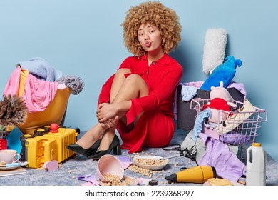 Romantic curly haired young lady in long red dress and black high heeled shoes sits on floor with clothes toys and food around poses in messy room sends air kiss isolated over blue background - Powered by Shutterstock