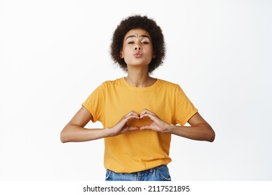 Romantic cute young african american woman pucker lips, reaching for kiss, showing I love you heart hand sign, standing in yellow tshirt over white background