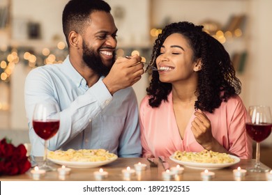 Romantic Date Concept. Smiling African American Guy Feeding His Cute Girlfriend With Pasta. Portrait Of Happy Black Family Sitting At Table And Eating Meal At Restaurant Or At Home, Drinking Red Wine