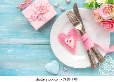 Romantic dinner concept. Table place setting with a white plate, vintage silverware tied with a pink ribbon and many different heart shape decorations, a gift and flowers With copy space - Powered by Shutterstock