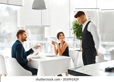 Romantic Dinner. Happy Smiling Couple In Love Reading Menu And Ordering Food To Waiter In Luxury Gourmet Restaurant. People Celebrating Anniversary Or Valentine's Day. Romance, Relationships Concept.