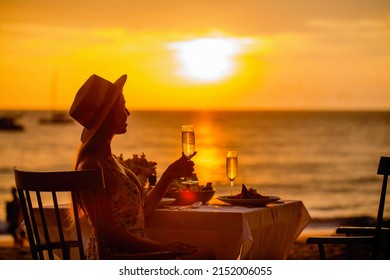 Romantic dinner near sea on sunset. Woman sitting alone, waiting husband on table set for a romantic meal on beach sky and ocean on background. Dinner for a couple in love in luxury outdoor restaurant