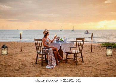 Romantic Dinner On Sunset. Woman Sitting Alone On Table Set With Lantern For A Romantic Meal On Beach, Yachts And Ocean On Background. Dinner For A Couple In Love In Luxury Outdoor Restaurant