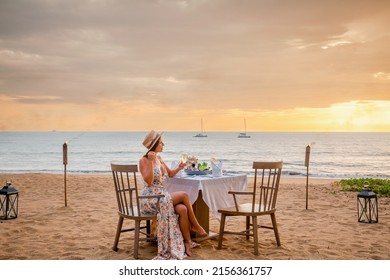 Romantic Dinner On Sunset. Woman Sitting Alone On Table Set With Lantern For A Romantic Meal On Beach, Yachts And Ocean On Background. Dinner For A Couple In Love In Luxury Outdoor Restaurant