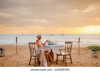 Romantic Dinner On Sunset. Woman Sitting Alone On Table Set With Lantern For A Romantic Meal On Beach, Yachts And Ocean On Background. Dinner For A Couple In Love In Luxury Outdoor Restaurant