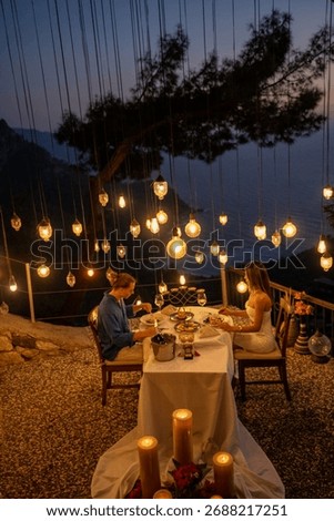 A romantic dinner table set for two, with a couple enjoying a sea view at sunset.

