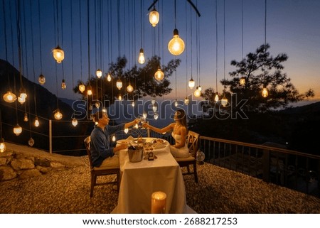 A romantic dinner table set for two, with a couple enjoying a sea view at sunset.


