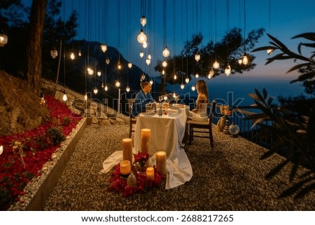 A romantic dinner table set for two, with a couple enjoying a sea view at sunset.

