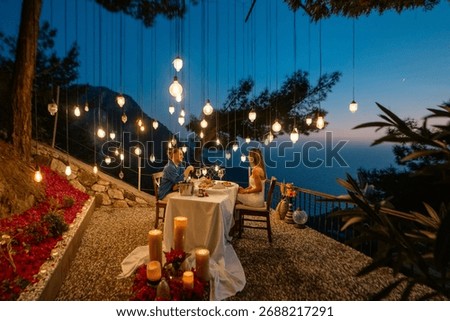 A romantic dinner table set for two, with a couple enjoying a sea view at sunset.

