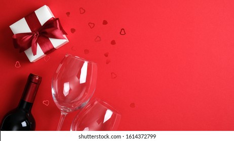 Romantic Dinner Table Setting For Valentine's Day. Bottle Of Wine, Glasses, Valentine Gift On Red Background. Top View With Copy Space