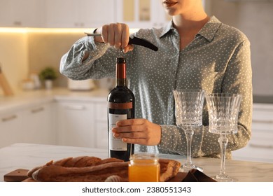 Romantic dinner. Woman opening wine bottle with corkscrew at table in kitchen, closeup