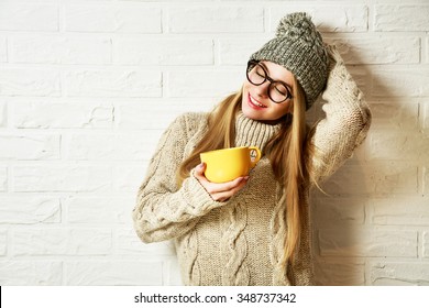 Romantic Dreaming Hipster Girl in Knitted Sweater and Beanie Hat with a Mug in Hands at White Brick Wall Background. Winter Warming Up Concept.