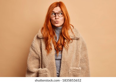 Romantic Dreamy Woman Keeps Lips Folded And Looks Away Imagines Something Pleasant Dressed In Fur Coat Wears Optical Glasses Isolated Over Beige Background. Indoor Studio Shot. Winter Outerwear