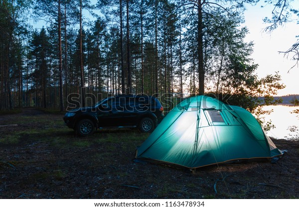 Romantic evening night landscape with a tent in the
forest near lake. The light from the lantern in a tent. Camping in
the wild nature. Car and portable table and chairs, green tourist
tent.
