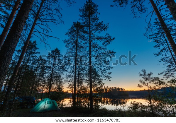 Romantic evening night landscape with a tent in the
forest near lake. The light from the lantern in a tent. Camping in
the wild nature. Car and portable table and chairs, green tourist
tent.