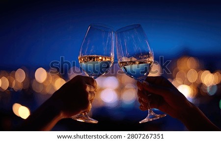 A romantic evening scene with two hands holding wine glasses filled with white wine, clinking them together in a toast. The background features a blurred cityscape with warm bokeh lights.