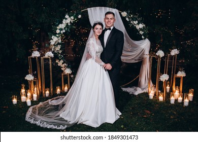 Romantic Evening Walk Of The Newlyweds Against The Backdrop Of A Flower Arch. Candlelight Ceremony.