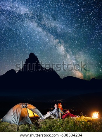 Romantic family hikers sitting by bonfire near lighting tent under incredibly beautiful starry sky. In the background silhouette of the mountains and luminous village in the valley at night. Low light