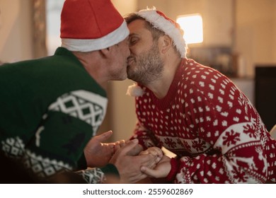 Romantic gay couple kissing and holding hands, wearing festive christmas sweaters and santa hats, celebrating the joy of the holiday season together at home, surrounded by warm decorations - Powered by Shutterstock