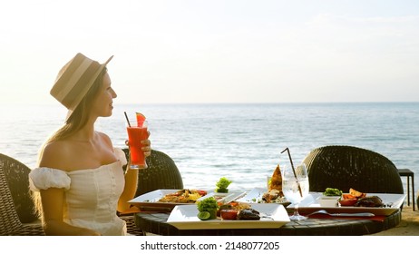 Romantic girl have lunch in tropical resort restaurant on beach, blue sea on background. Young woman in hat on vacation sip through paper straw healthy watermelon fruit cocktail in exotic hotel cafe.