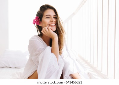 Romantic girl with sly smile in vintage blouse sitting on bed and touching her chin with hand. Portrait of dreamy cute young woman with flower in hairstyle resting in bedroom in morning