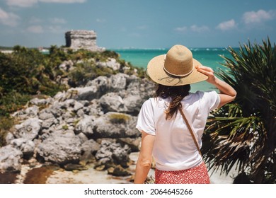 A romantic girl tourist in a hat stands on a cliff overlooking the Tulum ruins and the Caribbean Sea. Young woman view from the back against the backdrop of the ruins of Tulum, Mexico. Popular place