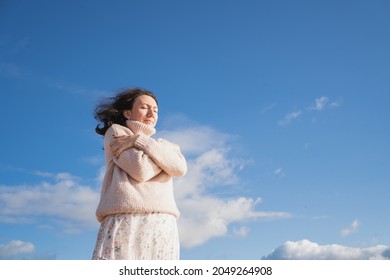 A romantic girl in a warm sweater hugs herself. The brunette dreamily closed her eyes. A woman stands against the sky with clouds.
